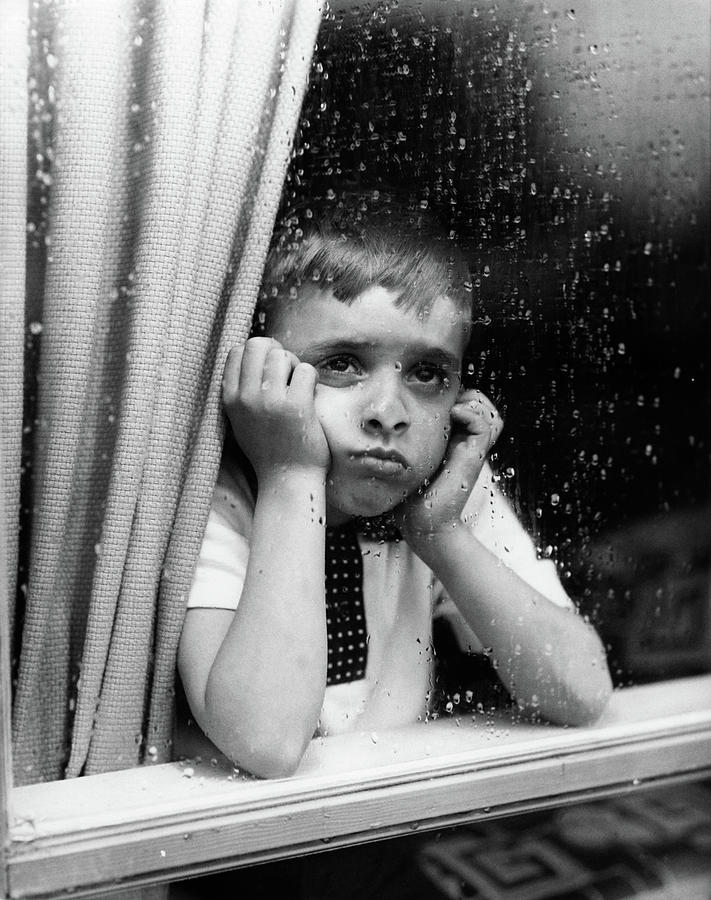 A sad boy looking out of a window with rain drops
